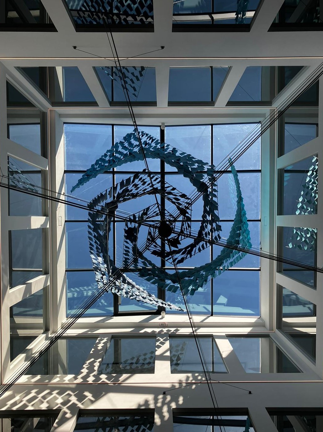 Atrium view of a large suspended vortex-shaped artwork at Solenis headquarters, designed by Twan Kerckhofs, composed of stainless steel rods and blue-hued paper leaflets