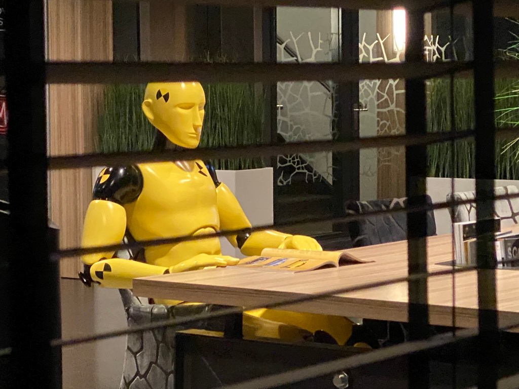 Life-size yellow crash test dummy named Theo sitting at a desk reading a magazine at Hedin Automotive headquarters, designed and produced by Art Nzo for Standis.