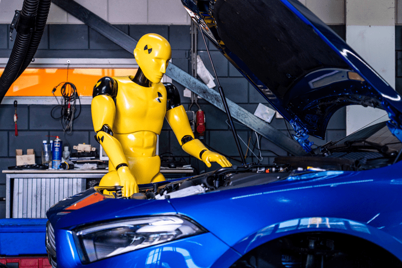 Yellow crash test dummy Theo repairing a blue car in a workshop, created by Art Nzo for Standis and Hedin Automotive recruitment campaigns. Photo courtesy of Hedin Automotive.